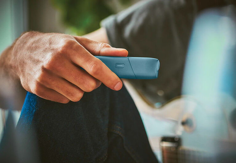 Turquoise IQOS Originals One device on a desk next to a tea cup.	 	 	