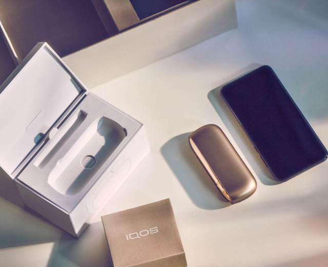 IQOS Originals Duo Holder and Pocket Charger in turquoise color on a desk, next to notebooks.	 	 	