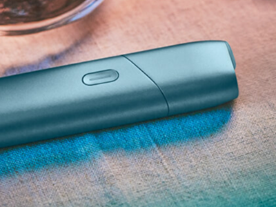 Turquoise IQOS Originals One device on a desk next to a tea cup.