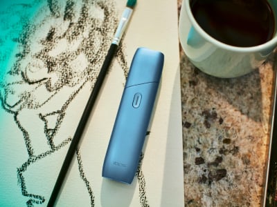 Turquoise IQOS Originals One device on a painting, next to a coffee cup.